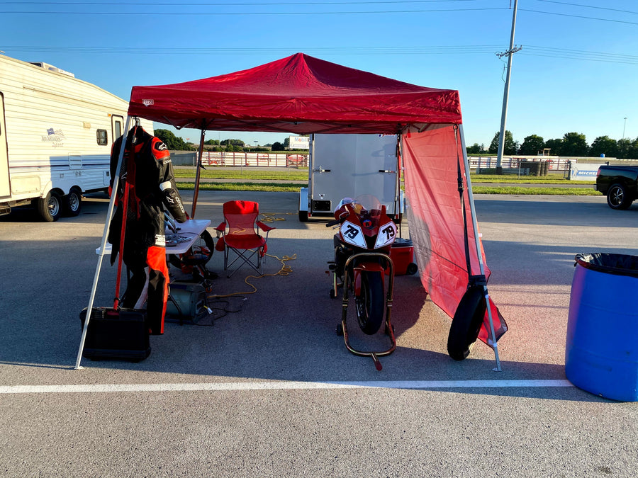 A simple track day paddock setup: red pop-up canopy over a folding table and camp chair, leathers hanging from a canopy leg, and a number-79 superbike on stands with a cooler and tool case alongside.