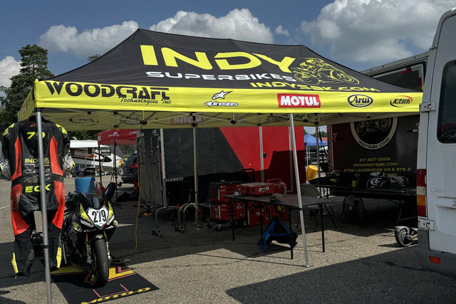 Indy Superbikes paddock setup at the track: a race suit hanging on the canopy leg, a number-279 superbike on stands, and tool chests under the awning.