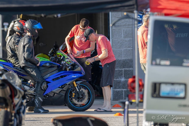Track day staff applying a tech inspection sticker to a blue Yamaha R6 while the geared-up rider sits on the bike in the paddock
